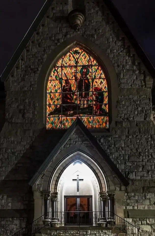 A nighttime photo of a stone church facade, featuring a brightly lit arched stained-glass window depicting religious figures…