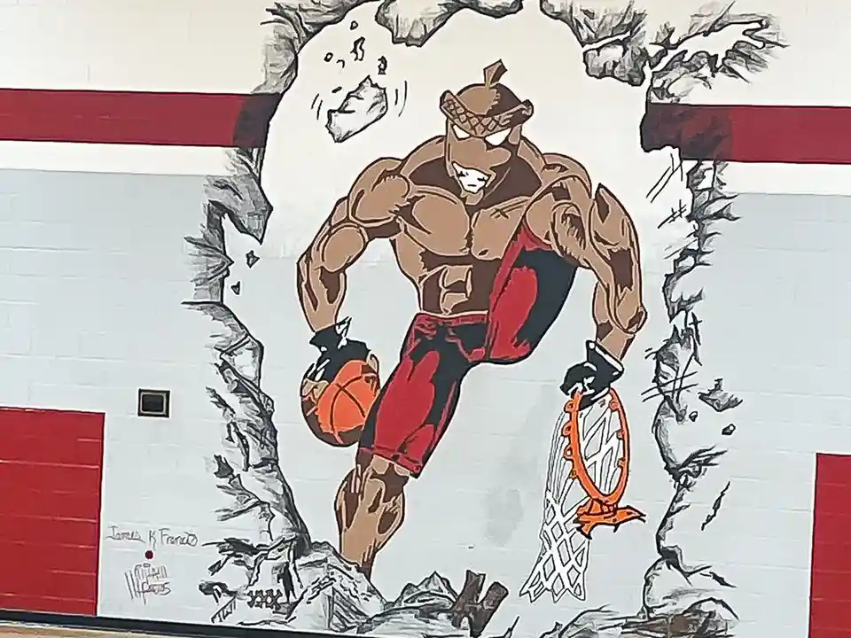 A mural on a white brick wall with red trim depicts a muscular, brown-skinned cartoon character wearing a helmet and red…