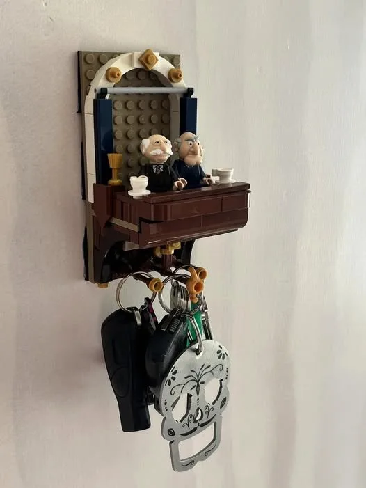 A LEGO key holder mounted on a wall features two LEGO minifigures resembling elderly men sitting at a small desk with…