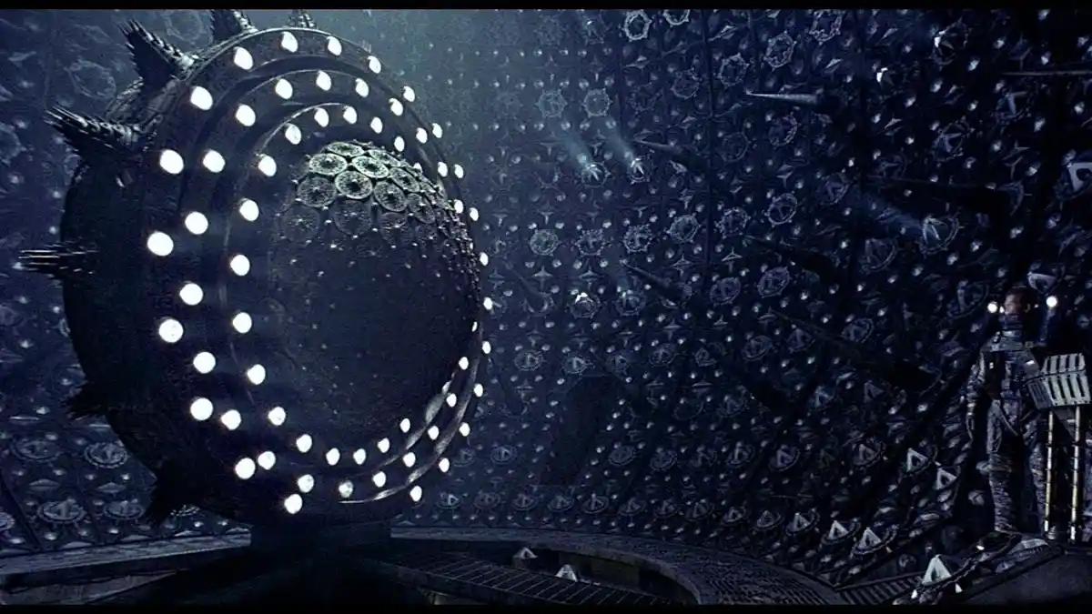 A person in a futuristic suit stands in a dark, ornate chamber with intricate patterns on the walls, looking at a large…