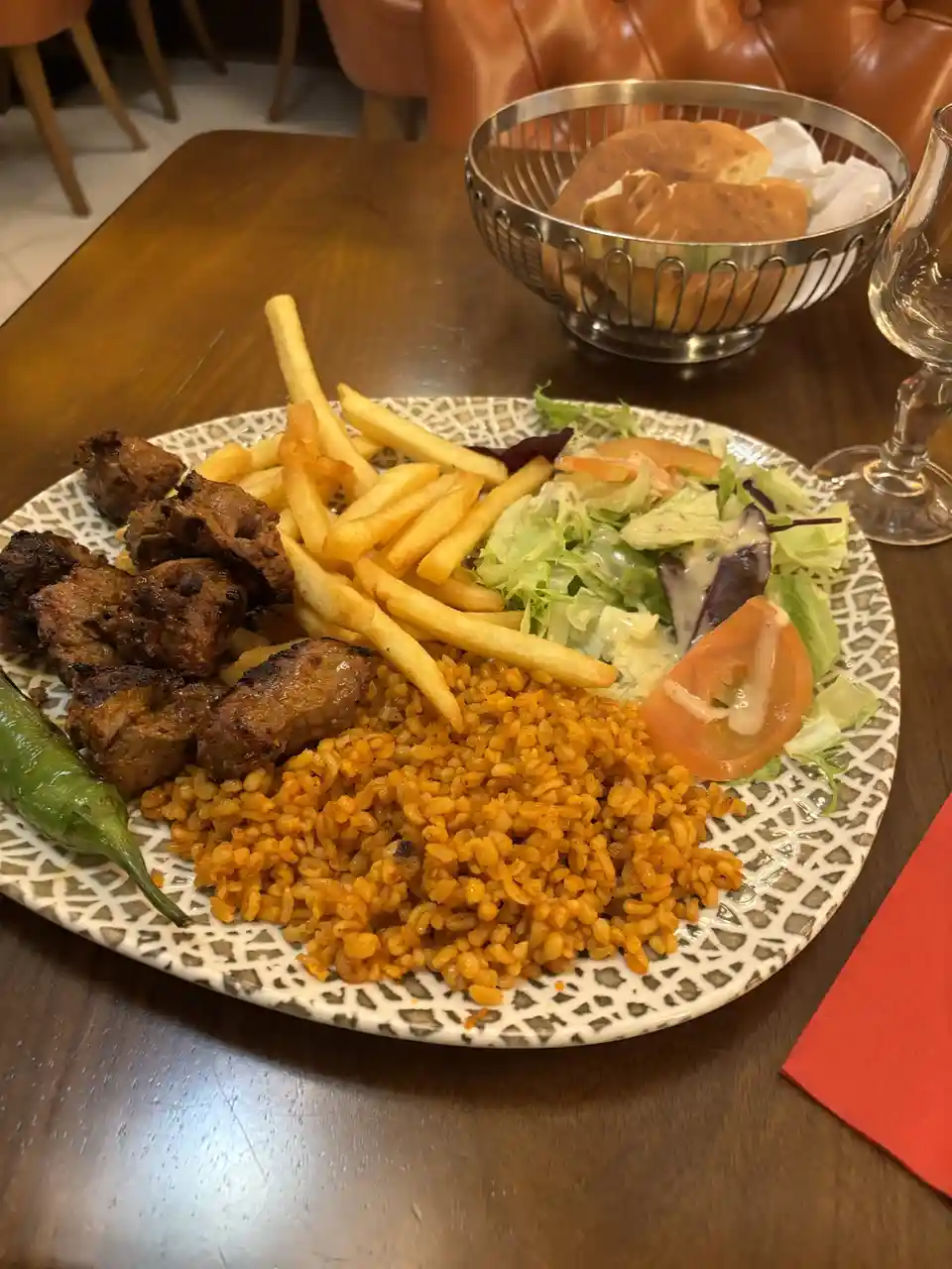 A plate of food featuring grilled meat, french fries, salad, and orange-colored rice sits on a wooden table. In the…