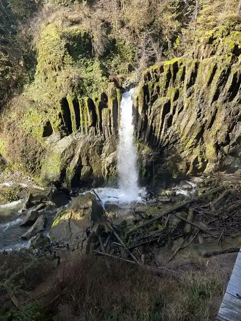 A scenic waterfall cascades down moss-covered, columnar basalt cliffs into a rocky stream below. Several small human figures…