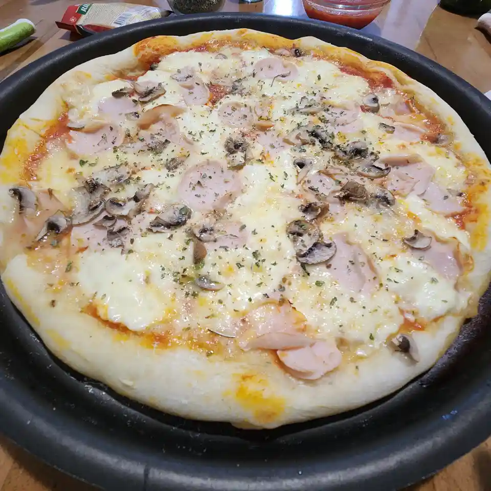 A freshly baked pizza in a black pan, topped with melted cheese, sliced ham, and mushrooms, garnished with herbs. The text…