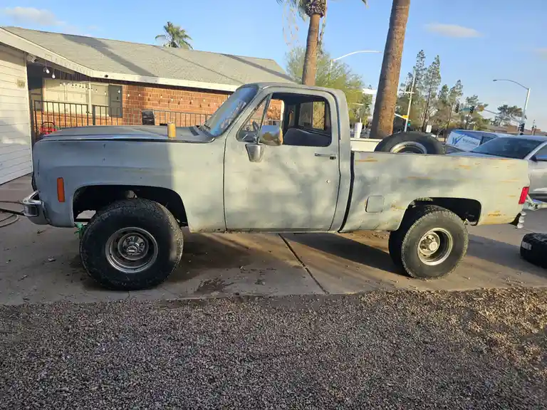 A weathered, light-gray vintage pickup truck with oversized tires is parked on a concrete driveway next to gravel, showing…