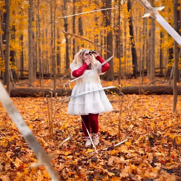 A person in a fantasy elf costume with pointed ears, wearing a white dress over red leggings and holding binoculars, stands…