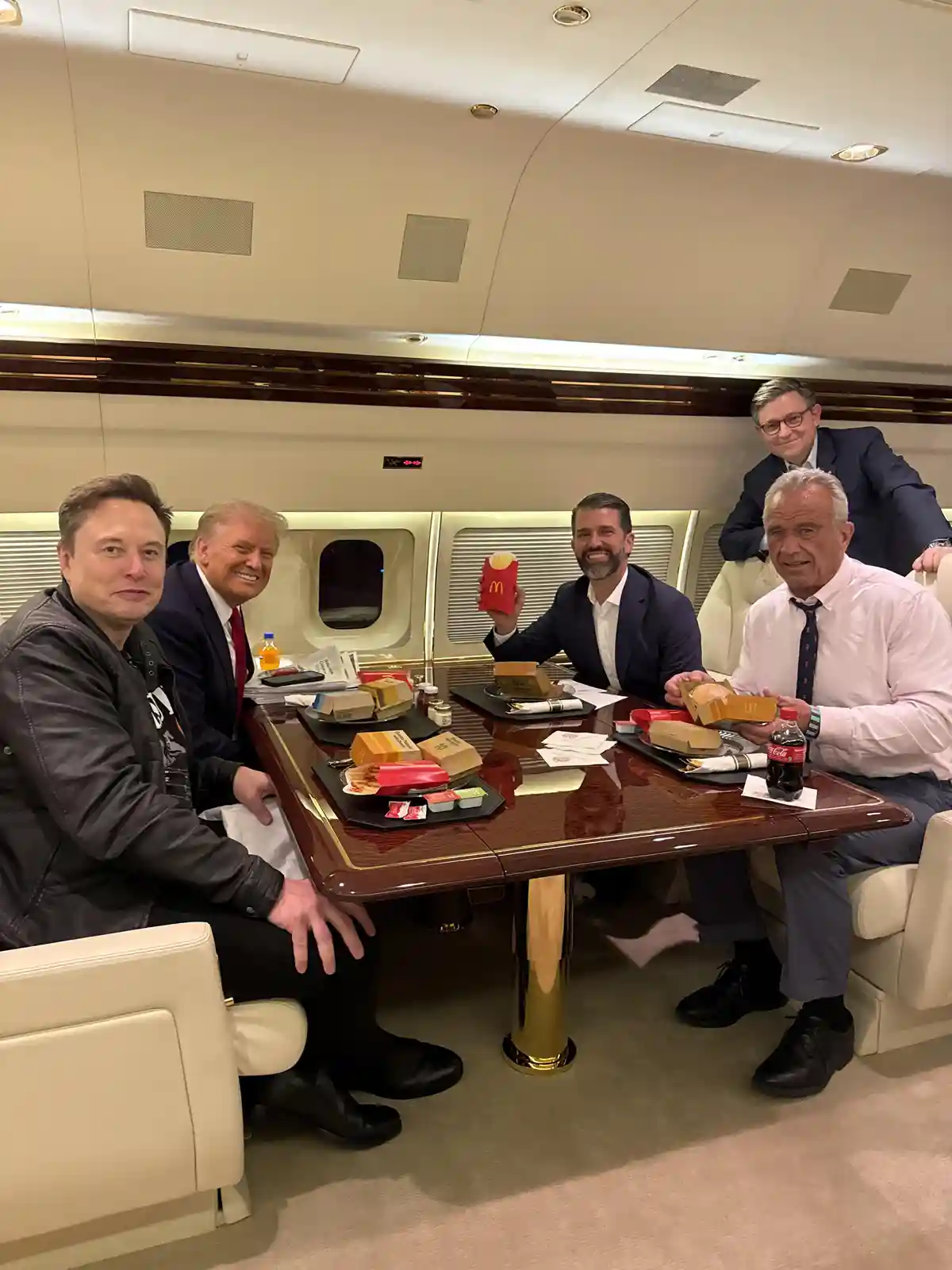 Five men are seated around a polished wooden table inside a luxurious private jet cabin, eating McDonald's food. Elon Musk…