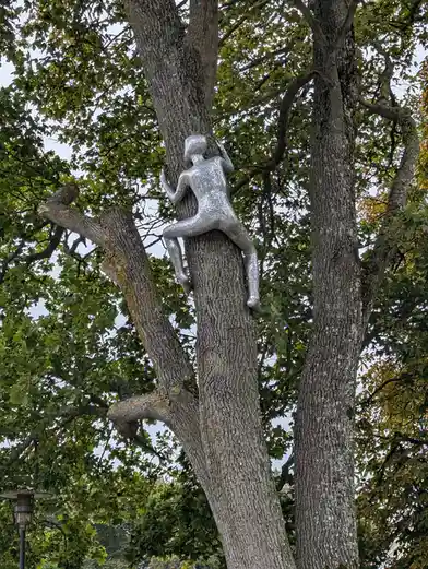 A silver, textured humanoid sculpture is affixed to the trunk of a large, leafy tree, appearing to climb upward. The…