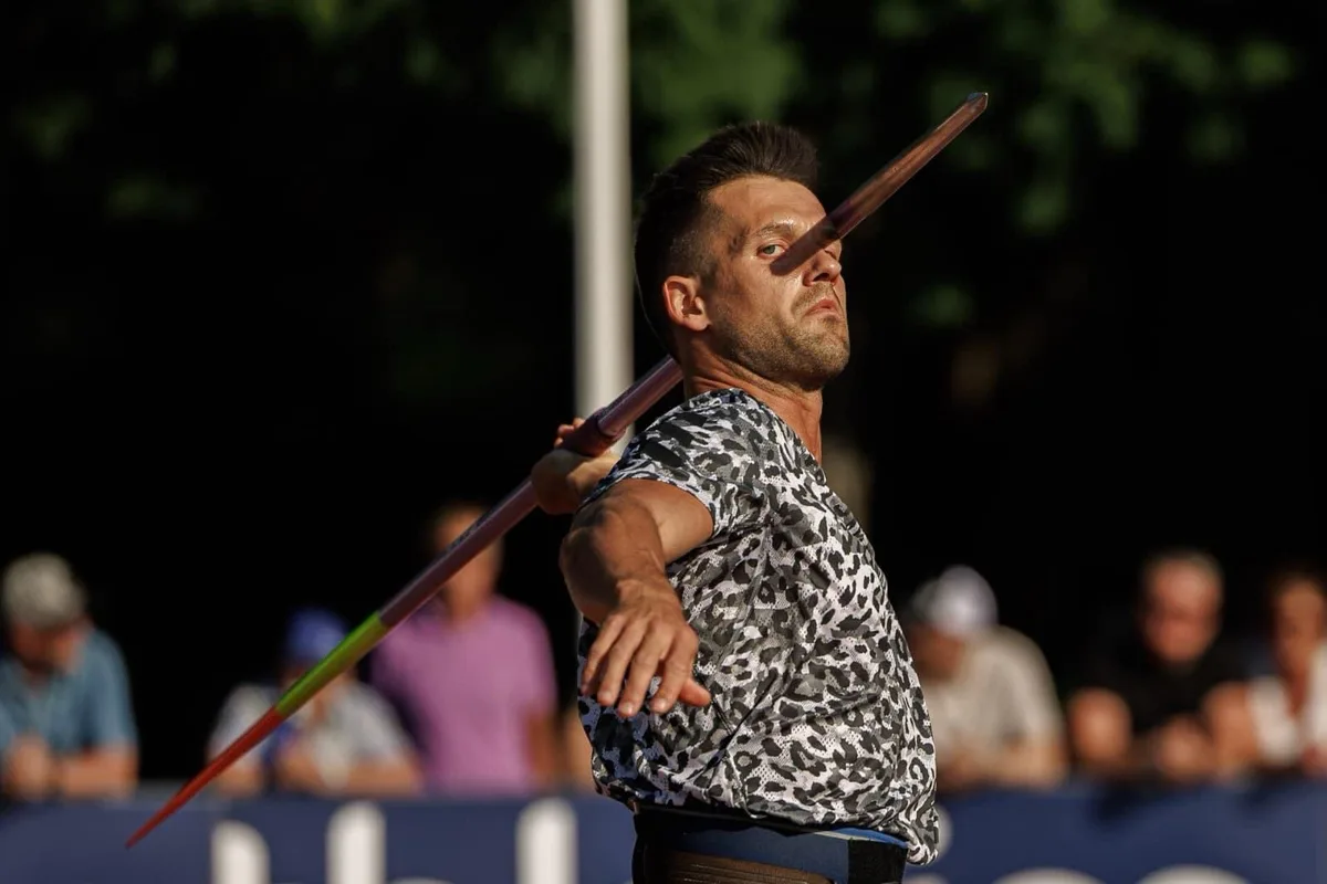 a male javelin thrower in a leopard-print shirt is captured mid-throw, with the javelin positioned horizontally across his…