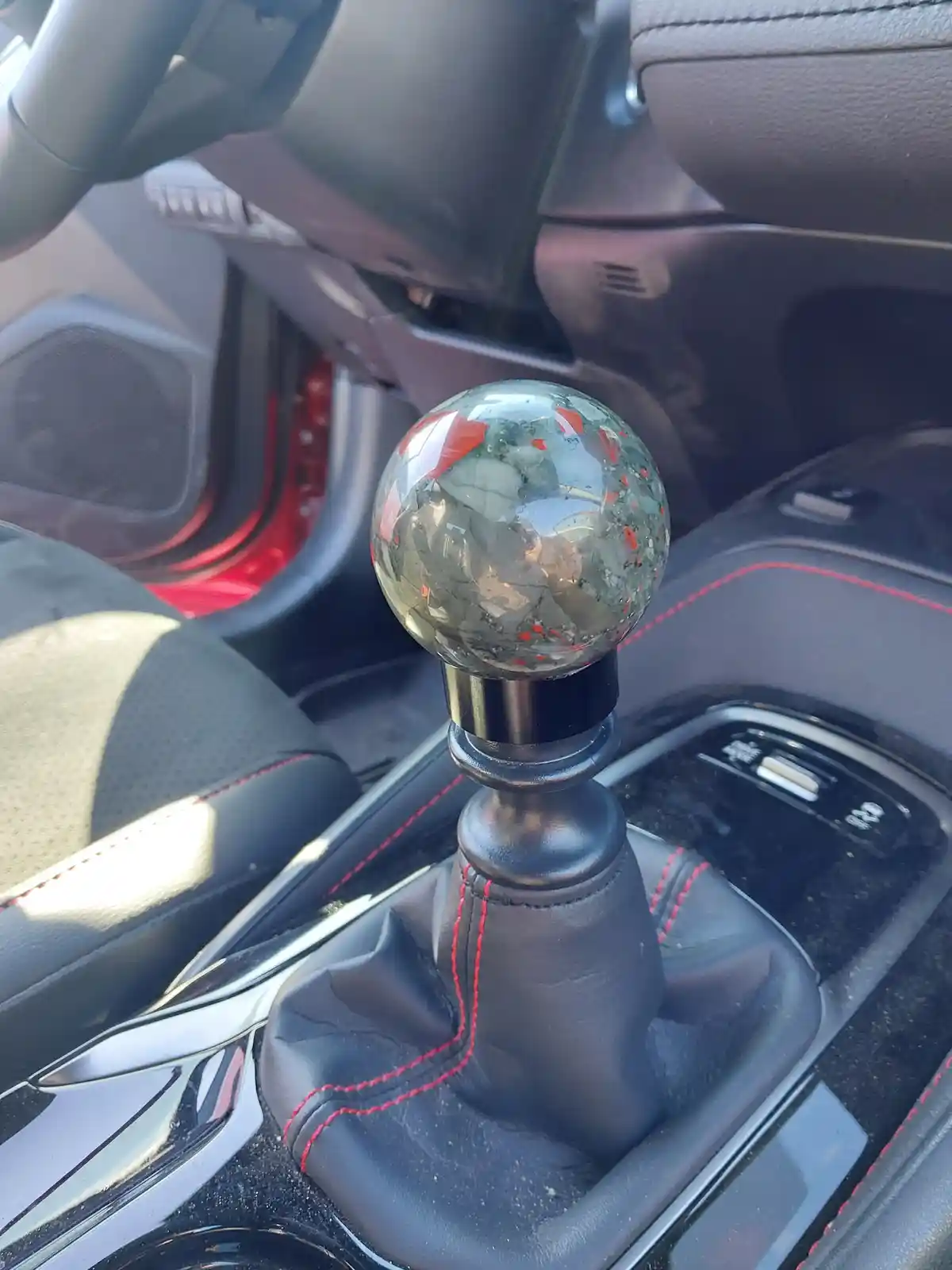 A close-up photo shows a manual car's gear shifter with a decorative, translucent sphere knob featuring red and gray swirls…