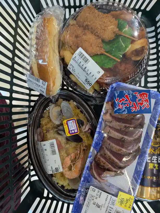 A black shopping basket holds four Japanese ready-to-eat food items: a bread roll, a fried food container, a seafood rice…
