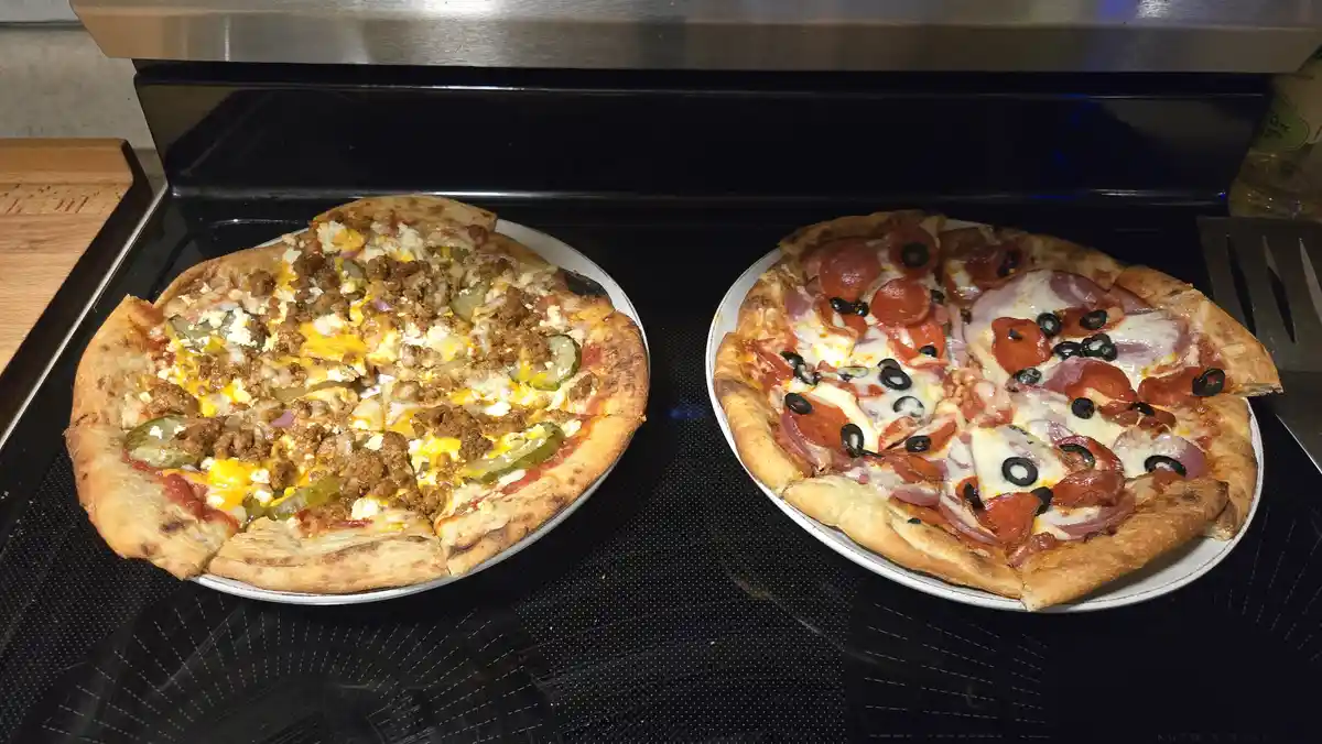 Two pizzas sit side-by-side on a black stovetop, each on a white plate. The pizza on the left has toppings like ground meat…