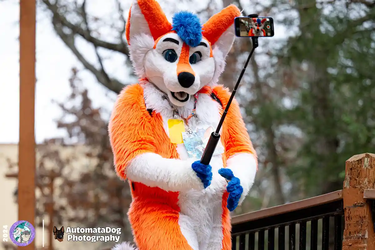 A person in a detailed orange and white fox fursuit with blue gloves and a blue pom-pom on their head is holding a selfie…