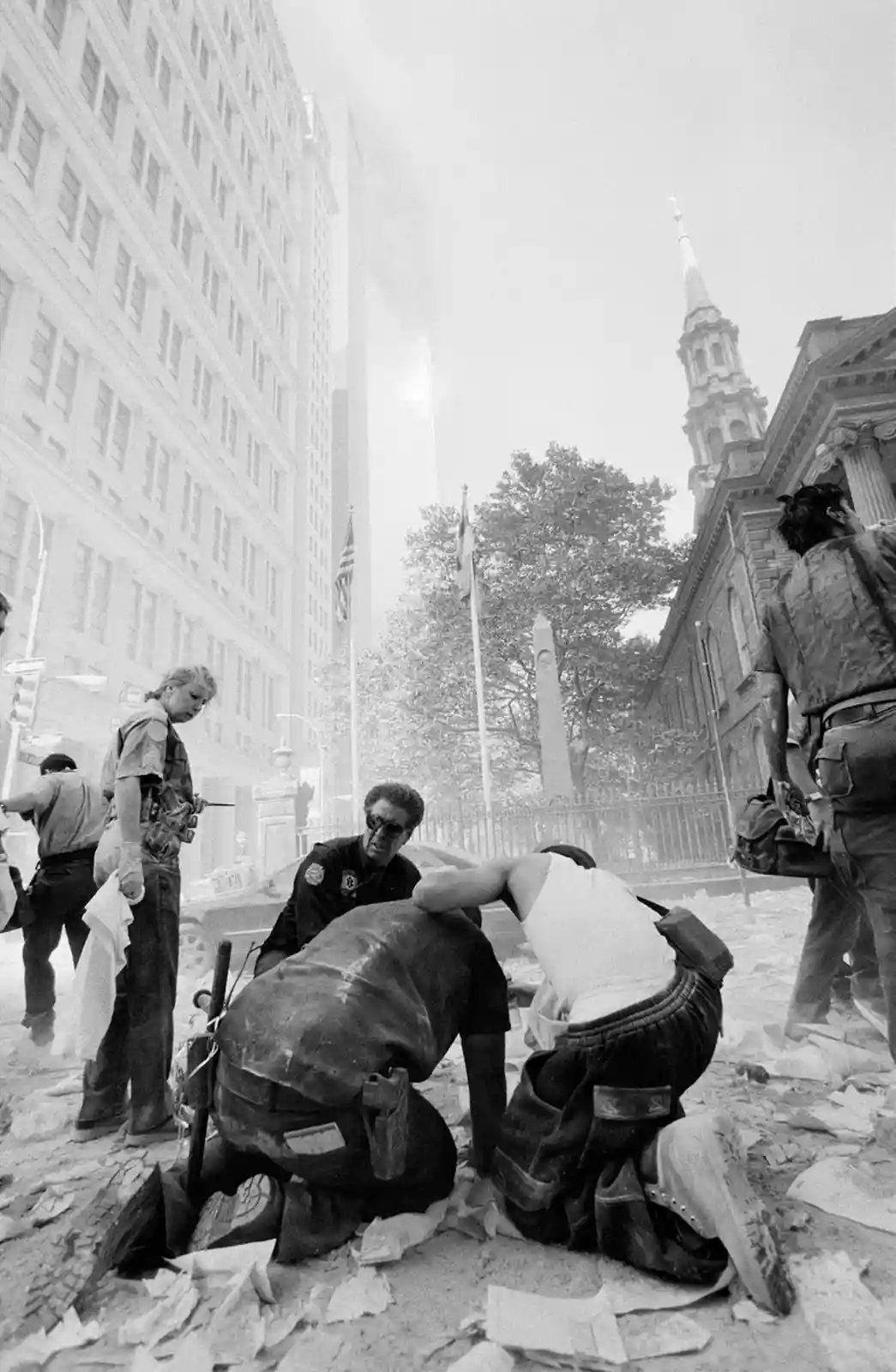 A black-and-white photo shows emergency responders and civilians amidst rubble and smoke in a city street, with a tall…