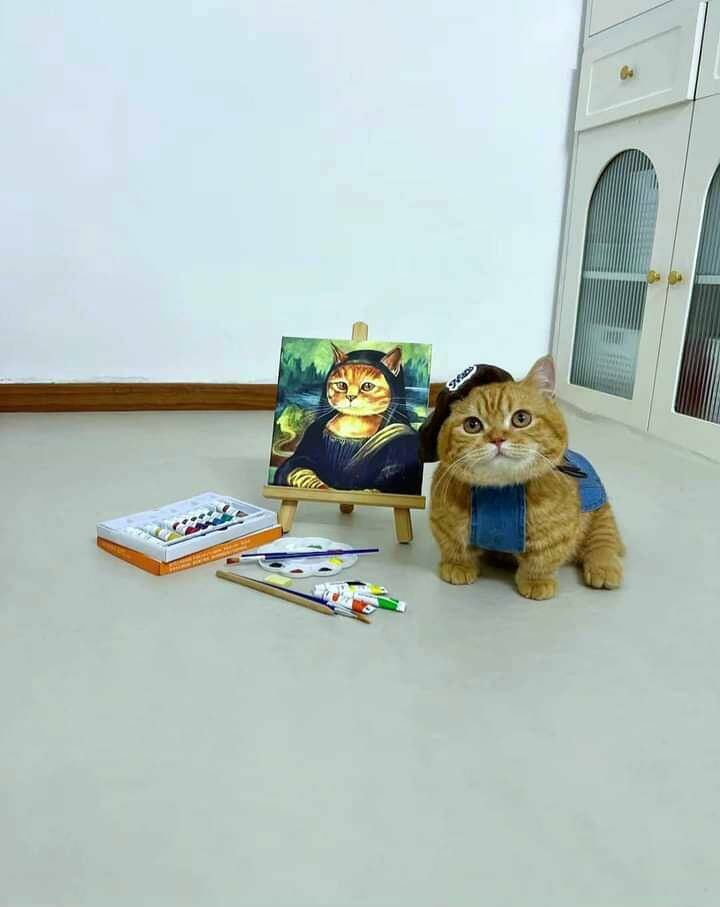 An orange tabby cat wearing a denim vest and a painter's cap sits on a floor next to an easel displaying a painting of…