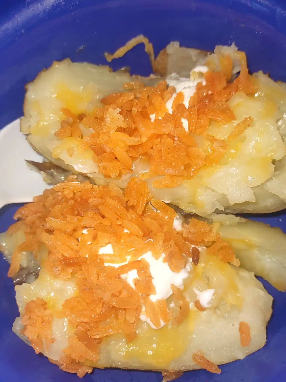 Two baked potato halves in a blue bowl, topped with melted cheese, sour cream, and crispy fried onions. The image has the…