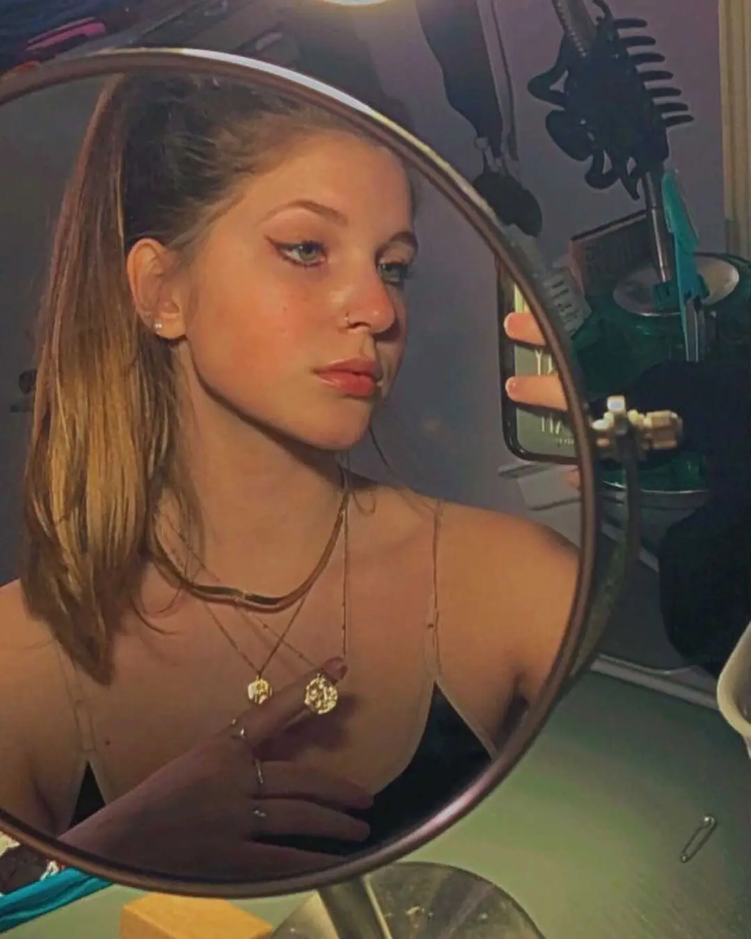 A young woman with light brown hair in a high ponytail looks at her reflection in a round vanity mirror while holding a…