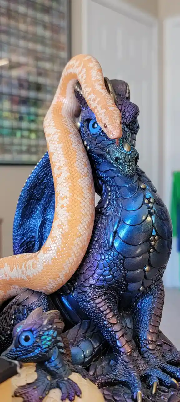 A live, peach-and-white patterned snake is coiled around a large, ornate blue and purple dragon statue, with its head…