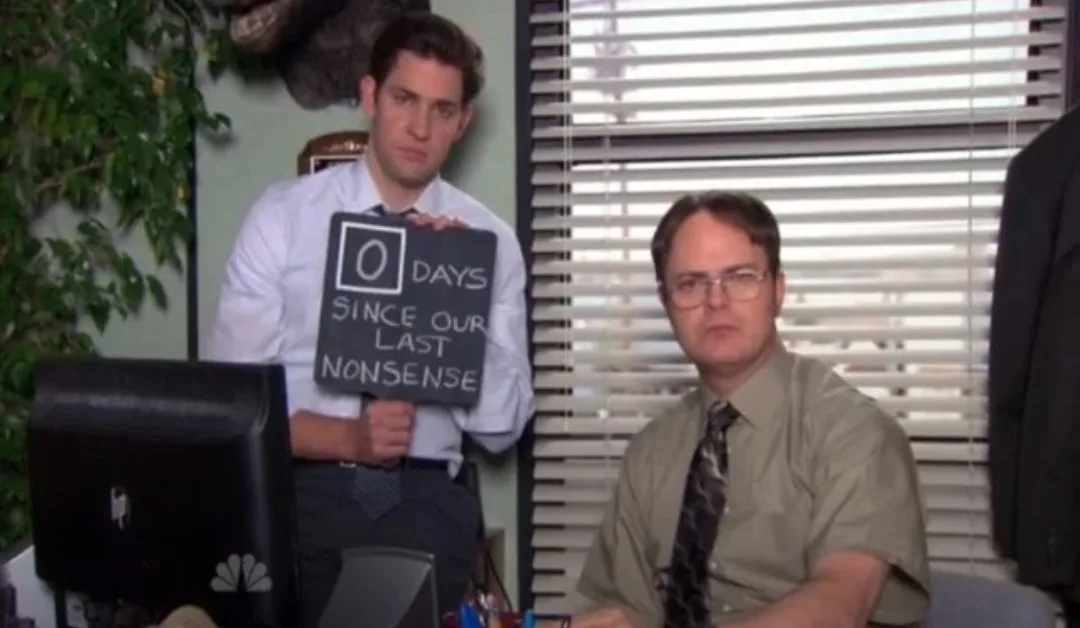 A man in a white shirt holds a chalkboard sign reading '0 DAYS SINCE OUR LAST NONSENSE' while standing behind another man in…