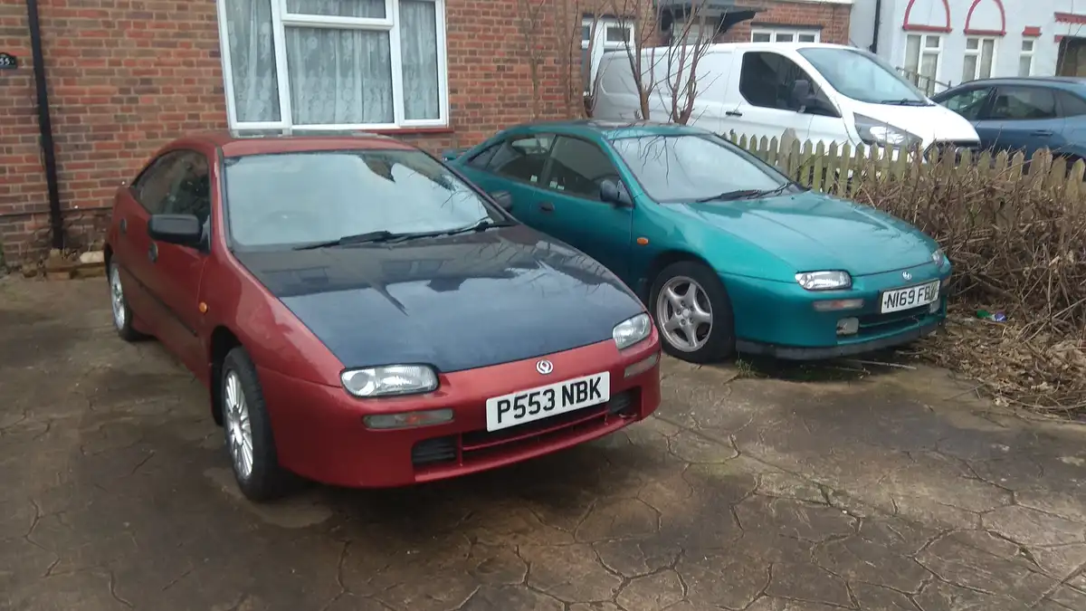 Two vintage cars, a red one with license plate P553 NBK and a teal one with license plate NI69 FBY, are parked side-by-side…
