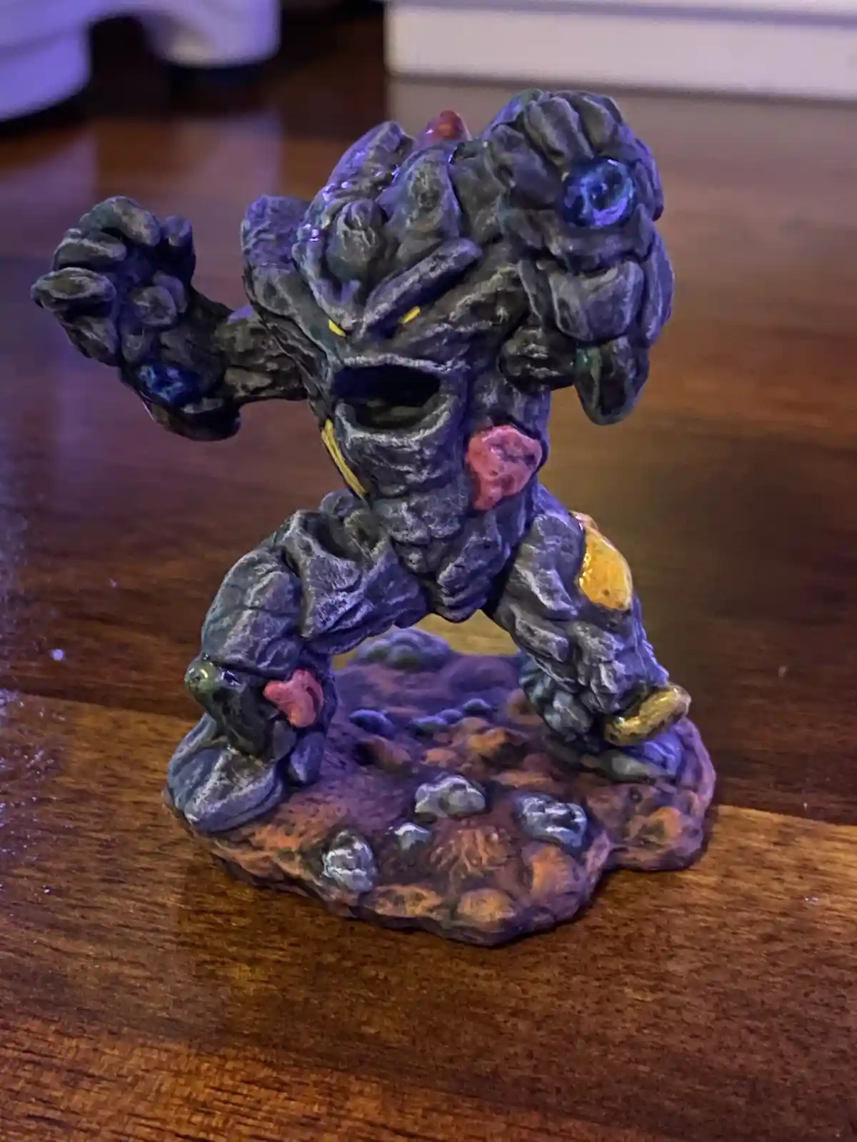 A small, stylized figurine of a rock-like creature with glowing eyes and colorful gem-like accents stands on a wooden…