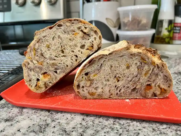 Two halves of a rustic, seeded bread loaf with visible nuts and orange flecks are displayed on a red cutting board in a…
