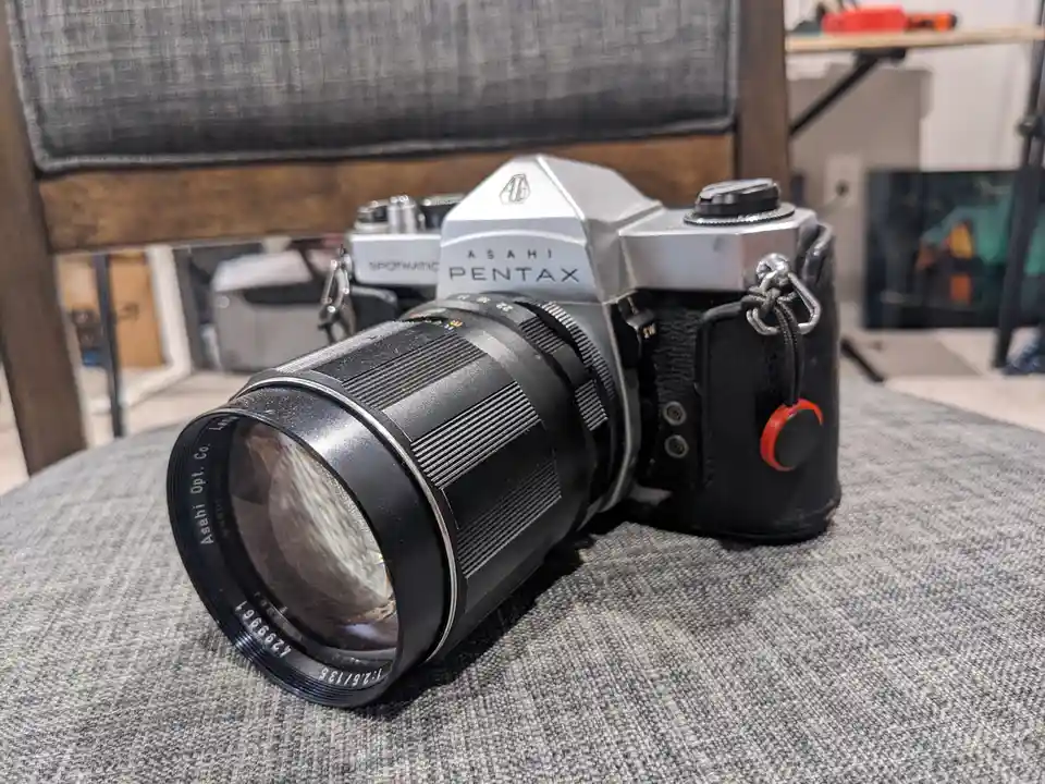 A silver Asahi Pentax film camera with a black lens and leather case rests on a gray fabric surface, with another camera…