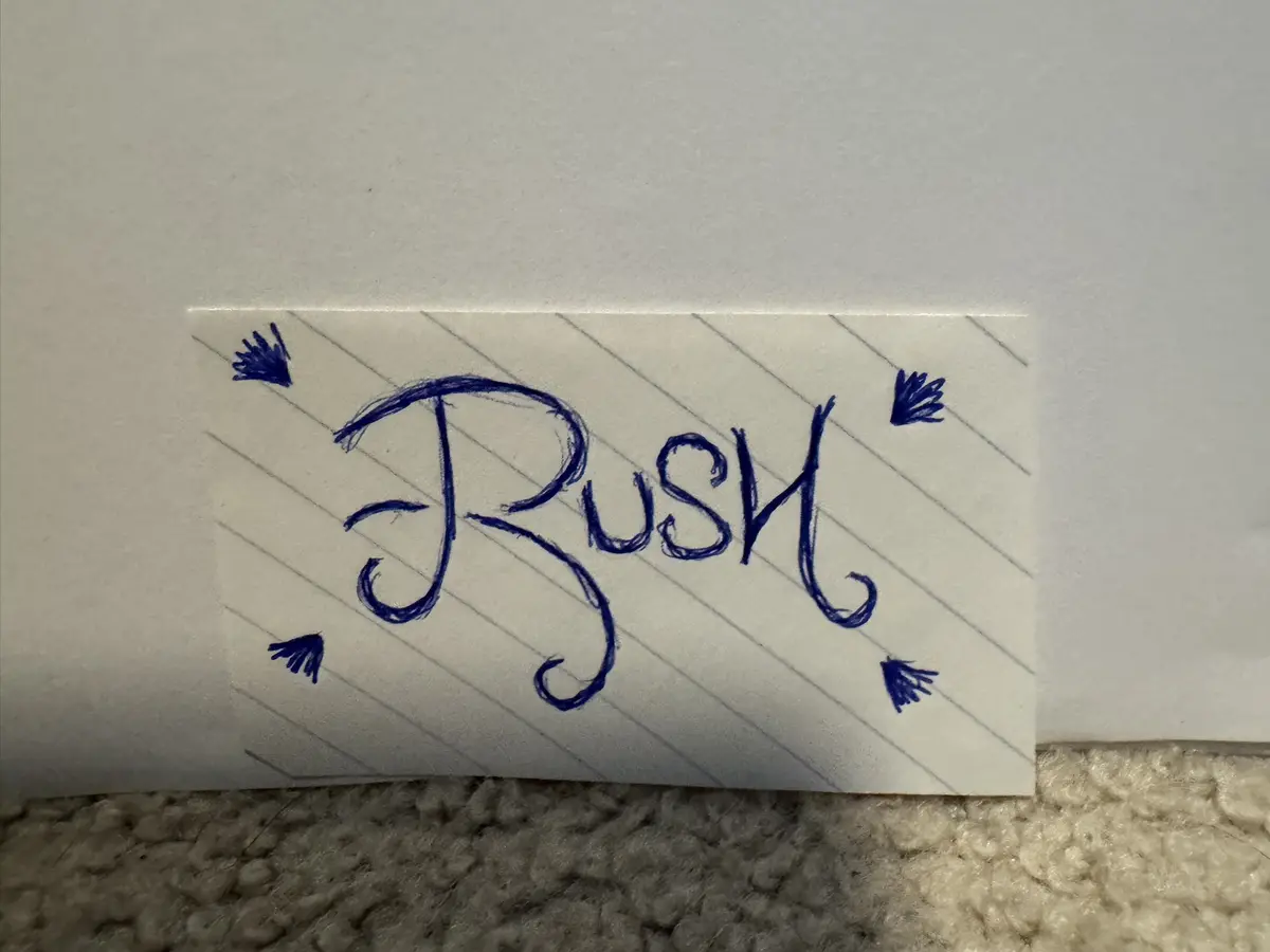 A close-up of a handwritten note on lined paper, written in blue ink, that says 'Rush' in a stylized cursive font. Four…