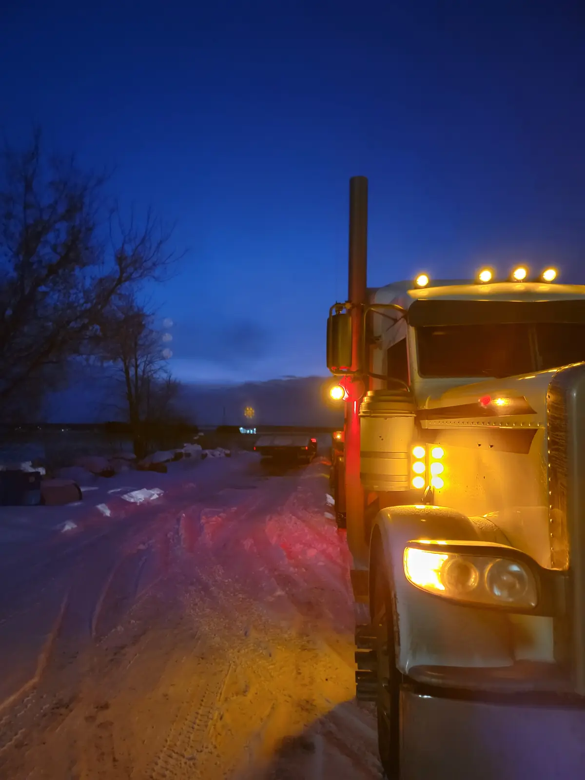 A white semi-truck with its headlights and roof lights on is parked on a snow-covered road at dusk. The truck's lights cast…