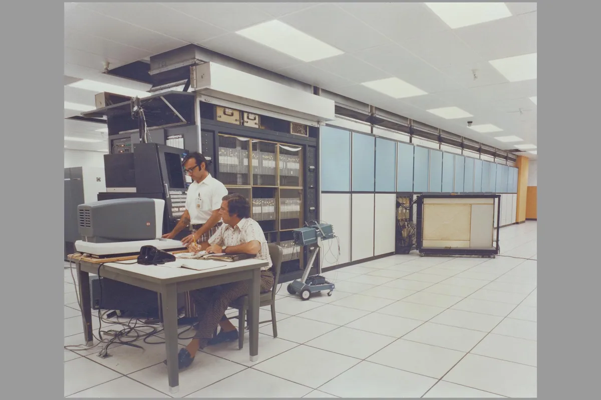 Two men are in a large, retro computer room with massive mainframe equipment. One man sits at a desk with an old CRT…