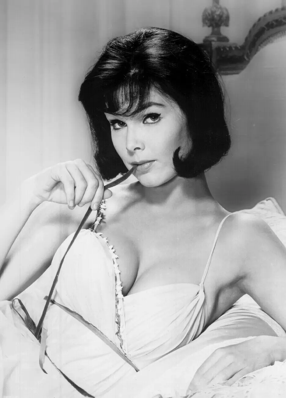 A black-and-white photo shows a woman with dark hair and a white dress holding a thin object to her lips while looking…