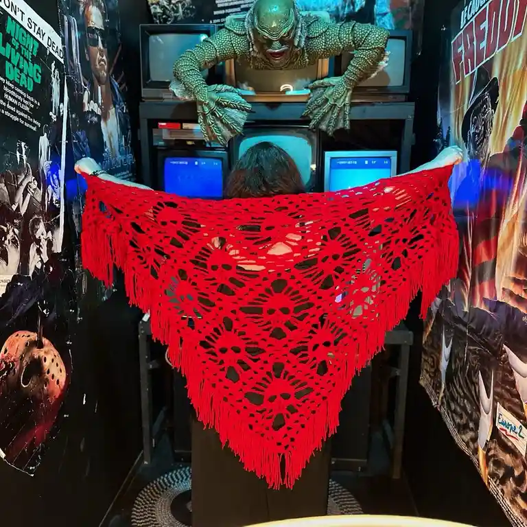 A person viewed from behind holds up a large, bright red crocheted shawl with intricate patterns and fringes, standing in…