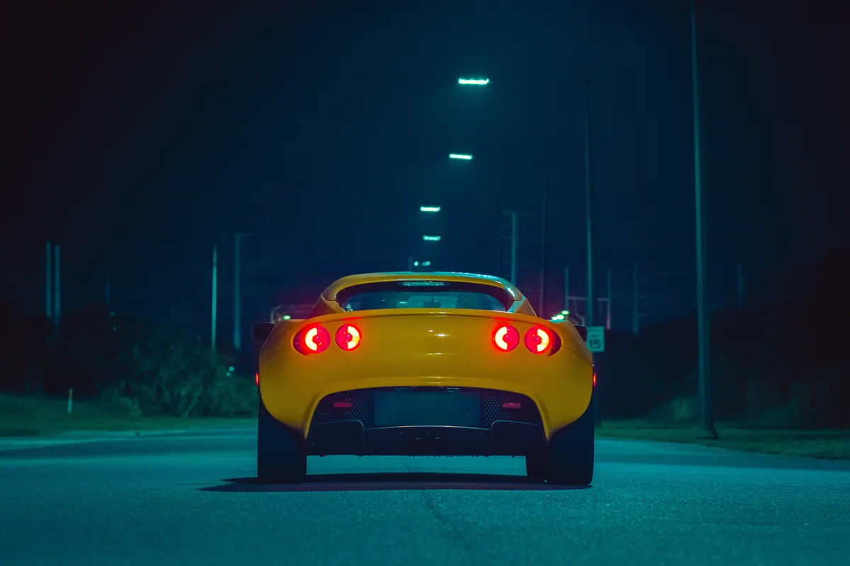 A yellow sports car is seen from behind on a dark, empty road at night, its distinctive circular red taillights glowing…