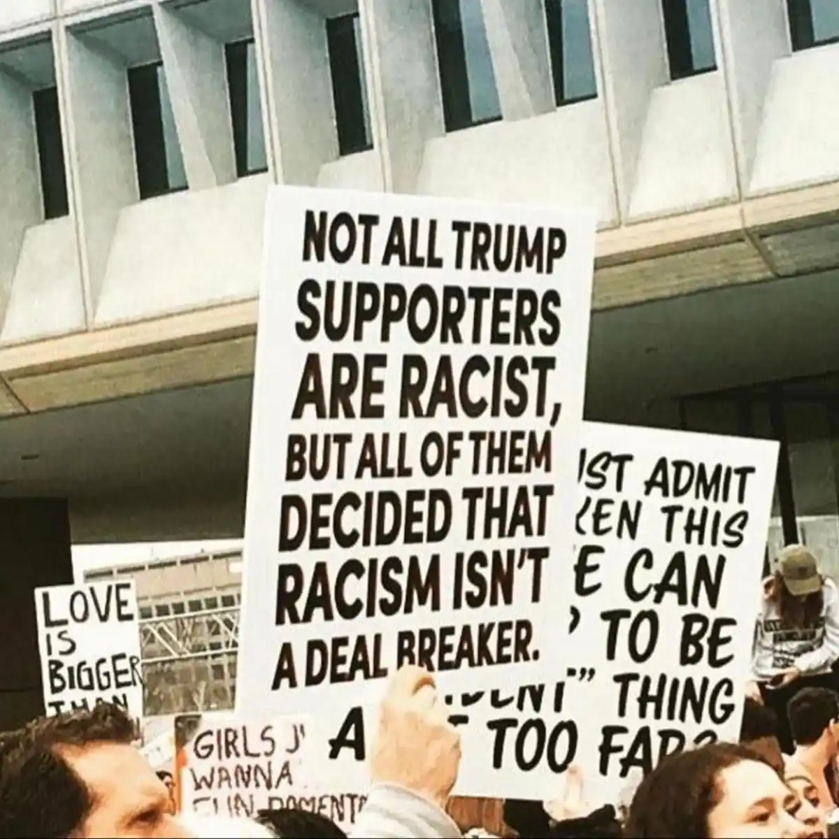 A protest photo shows multiple signs held by demonstrators in front of a modern building. The most prominent sign reads 'NOT…