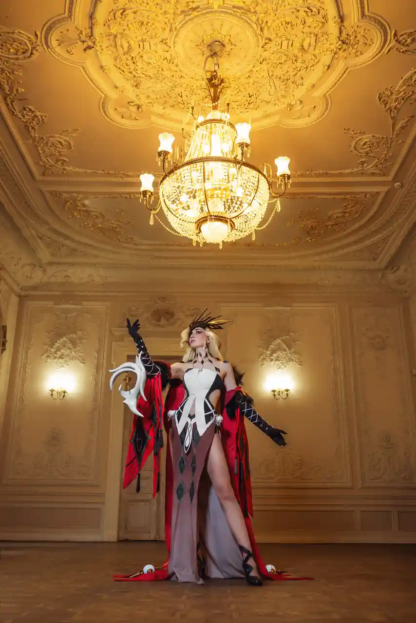 A cosplayer in an elaborate black, white, and red costume with a feathered headdress and a large white horned prop stands in…