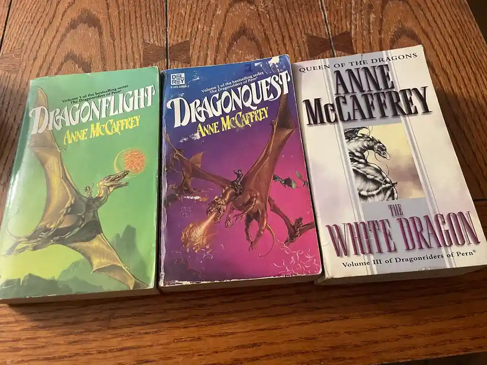 Three vintage paperback books by Anne McCaffrey from 'The Dragonriders of Pern' series are arranged side-by-side on a wooden…