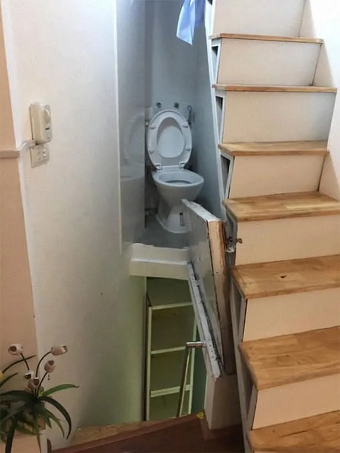 A white toilet is installed in a very narrow, cramped space directly under a wooden staircase, with a ladder visible below…