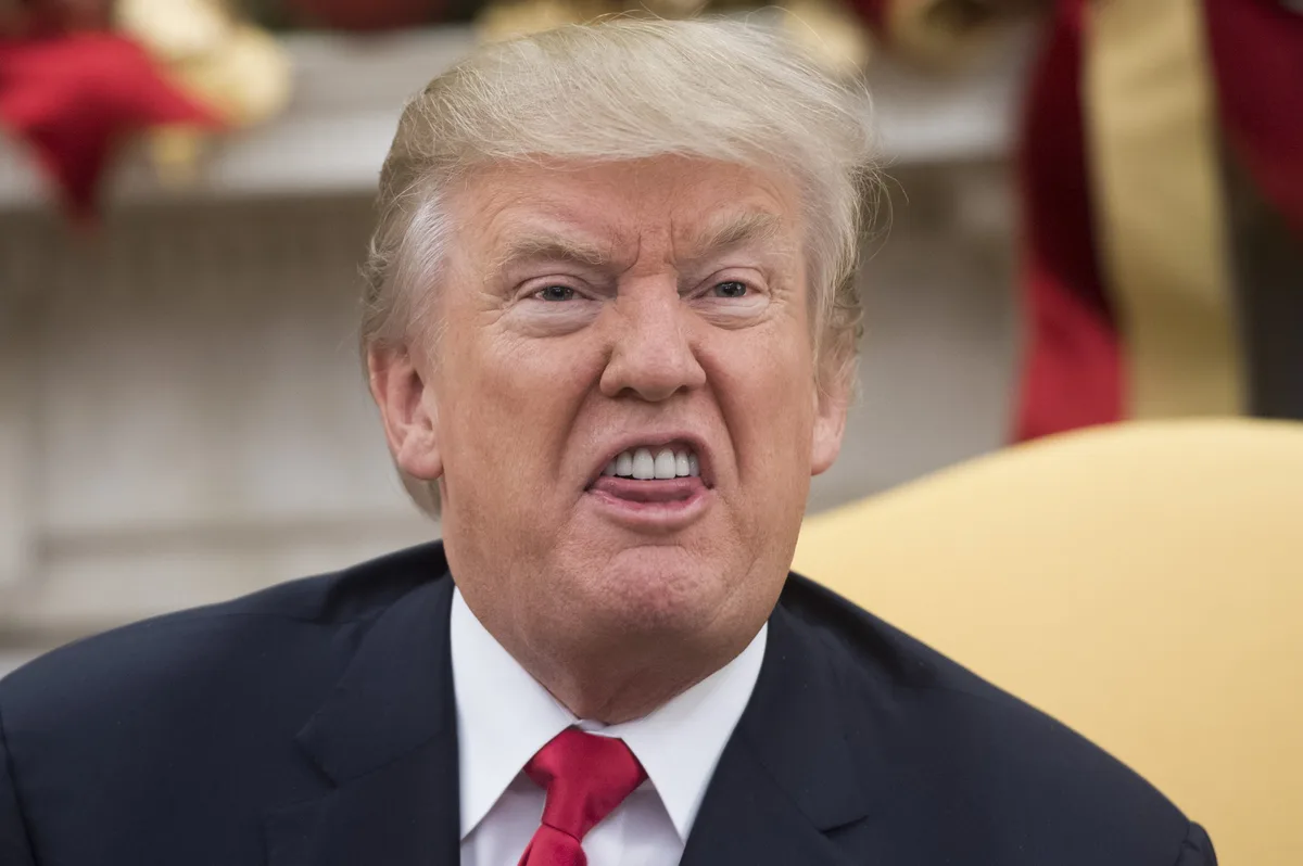 A close-up photo of Donald Trump with a furrowed brow and an angry, shouting expression, wearing a dark suit and red tie…