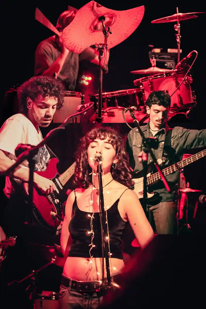 A band performs on stage under red lighting, with a female singer in the foreground wearing a black top and string lights…