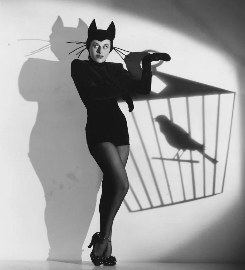 A woman in a black cat costume with ears and whiskers stands in a spotlight, her shadow on the wall forming a cat silhouette…