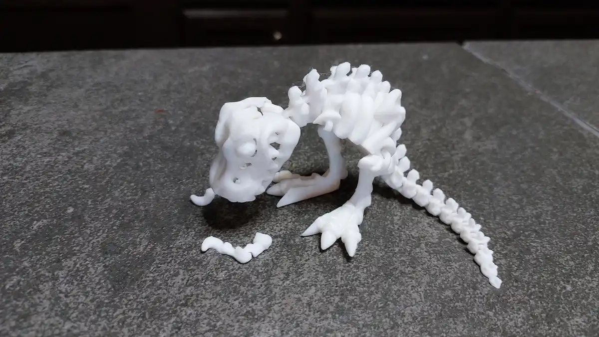 A white 3D-printed skeleton of a small reptile-like creature, possibly a lizard or gecko, is posed on a gray speckled…