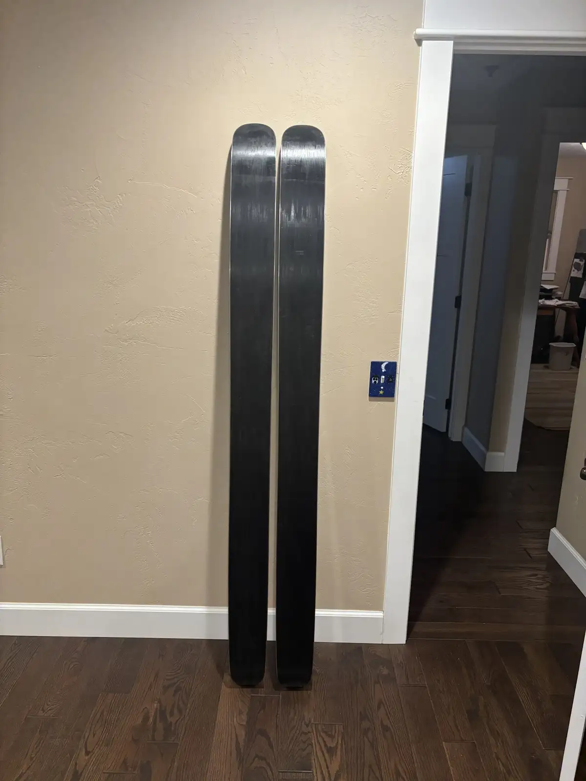 Two black skis stand vertically against a beige textured wall on a dark wood floor, next to a doorway. The overlaid text…