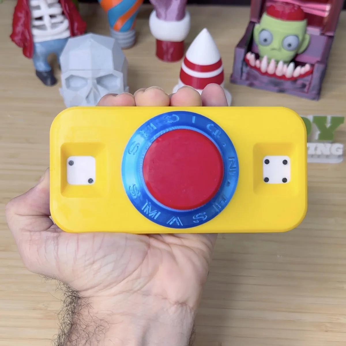 A hand holds a yellow 3D-printed toy resembling a camera with a large red button and a blue directional pad labeled with…