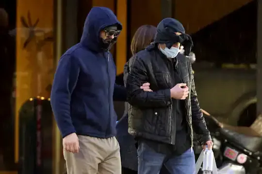 Two men walking outdoors in rainy weather, both wearing hooded jackets and face coverings. The man on the left wears a dark…
