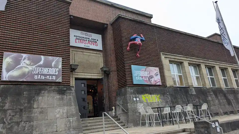 A brick building with a large Spider-Man statue clinging to its side, flanked by two 'Superheroes' exhibition posters. A…