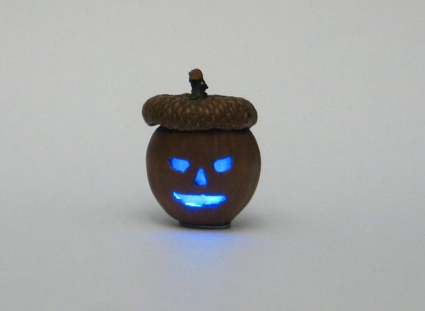 a small acorn has been carved into a jack-o-lantern face with glowing blue eyes and mouth, set against a plain white…