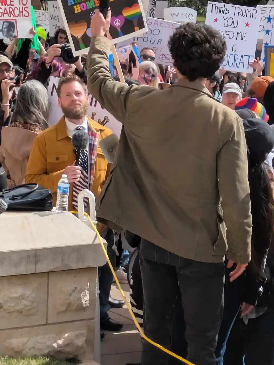 A man with curly hair, seen from behind, holds a sign at a protest while a reporter in a yellow jacket holds a microphone…