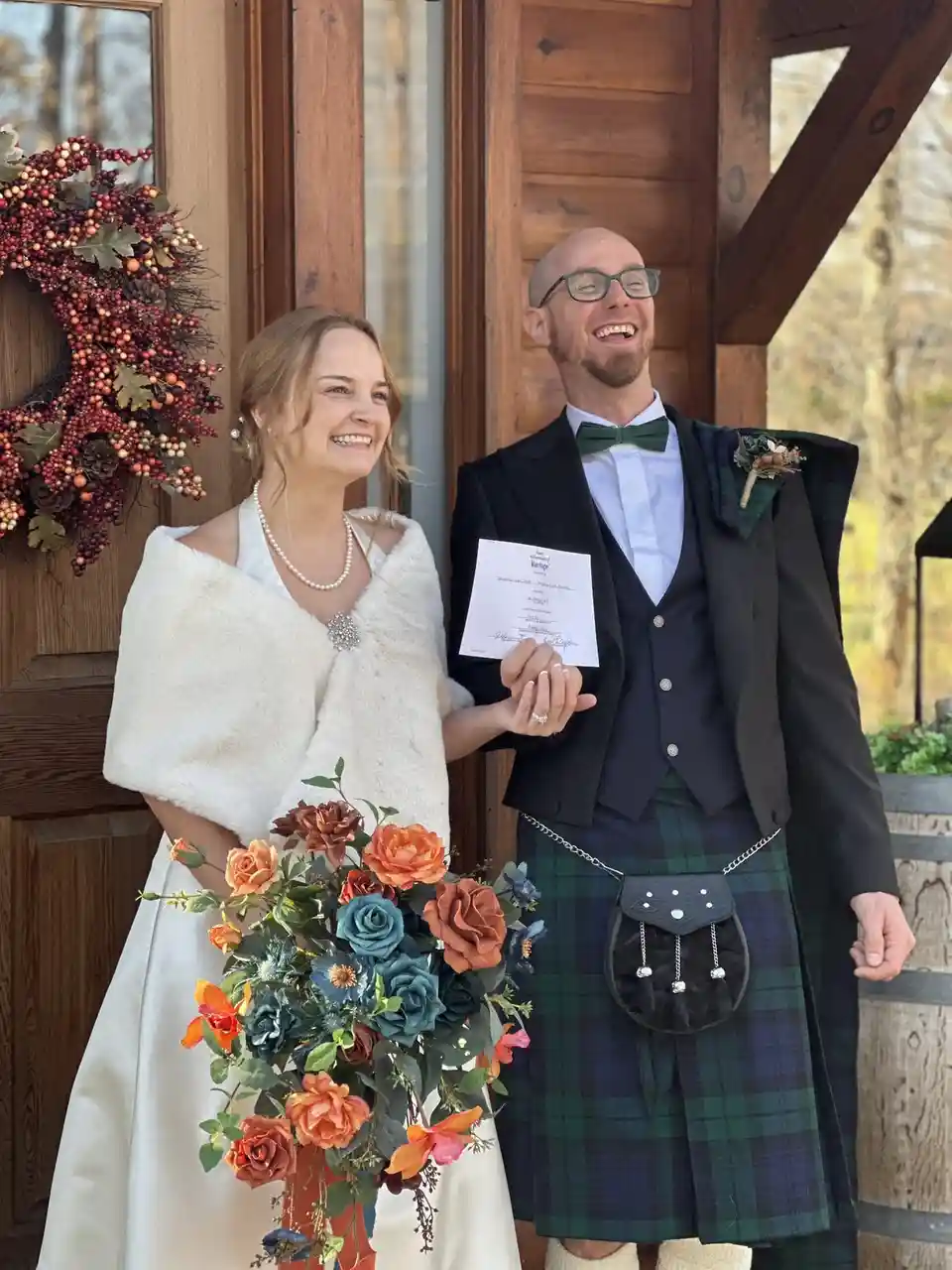 A smiling bride in a white dress and fur shawl holding a colorful bouquet stands beside a bald man in a green kilt, vest…