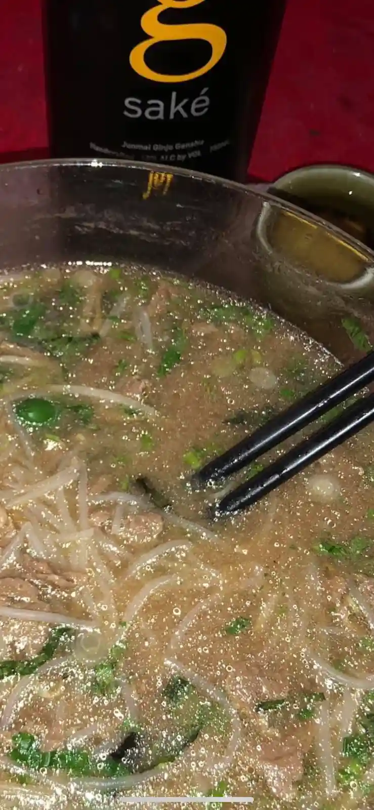 A close-up photo shows a steaming bowl of noodle soup with chopsticks resting in it, next to a bottle labeled 'sake'. The…