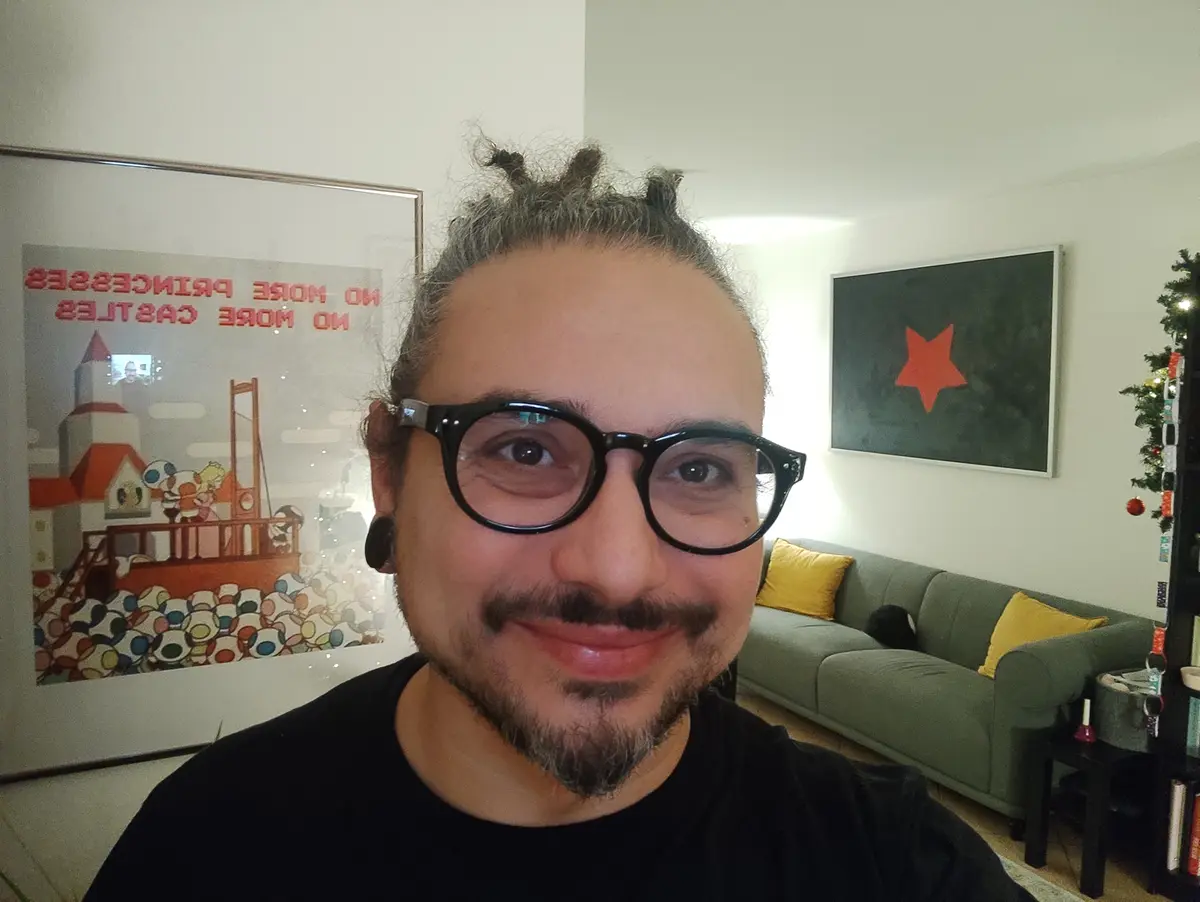 A man with glasses and a beard smiles at the camera in a living room. Behind him, a framed poster reads 'NO MORE PRINCESSES…