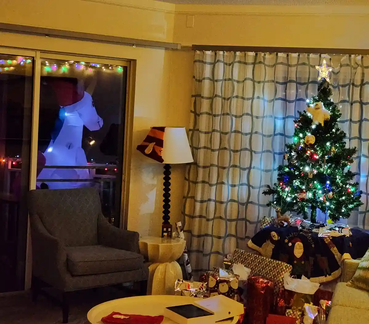 A cozy, warmly lit living room decorated for Christmas with a tree, presents, and a lamp, while an inflatable unicorn…